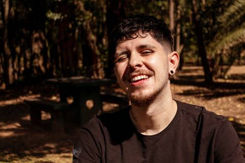 Matheus com árvores ao seu redor no parque do Ibirapuera, na capital de são paulo, sorrindo para a câmera, piscando de um olho.'