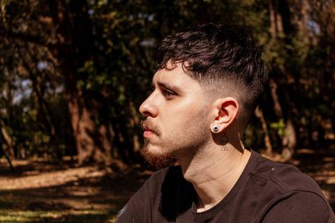 Matheus com árvores ao seu redor no parque do Ibirapuera, na capital de são paulo, lateral do rosto, sério.'