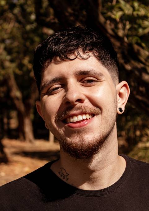 Matheus com árvores ao seu redor no parque do Ibirapuera, na capital de são paulo, enquadramento fechado em seu rosto, sorrindo para a câmera, frontal.'