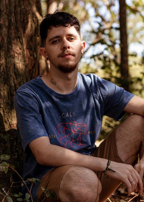 Matheus com árvores ao seu redor no parque do Ibirapuera, na capital de são paulo, encostado em árvore, olhando diretamente para a câmera.'