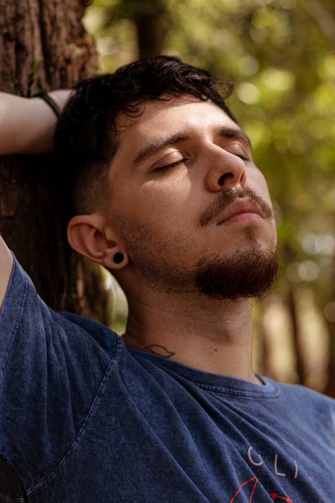 Matheus com árvores ao seu redor no parque do Ibirapuera, na capital de são paulo, encostado em árvore, olhos fechados e cabeça inclinada para o alto.'