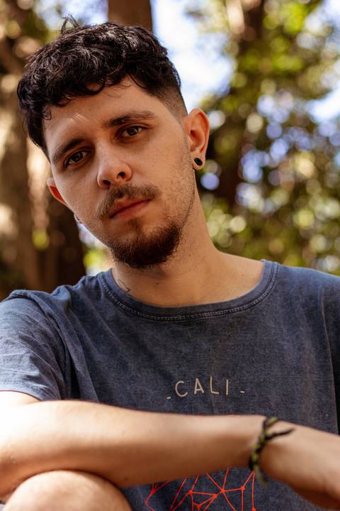 Matheus com árvores ao seu redor no parque do Ibirapuera, na capital de são paulo, olhando diretamente para mim, sério.'