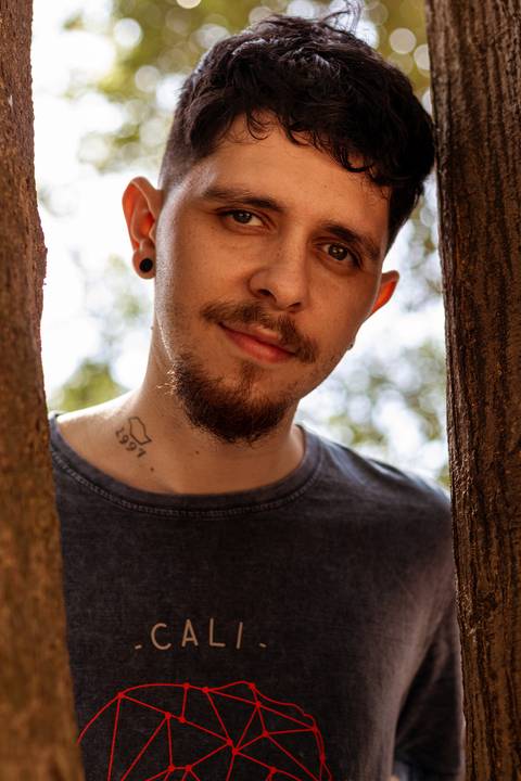 Matheus com árvores ao seu redor no parque do Ibirapuera, na capital de são paulo, entre duas árvores, sorrido leve.'