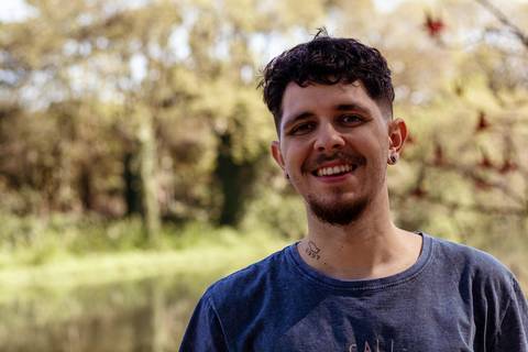 Matheus com árvores ao seu redor no parque do Ibirapuera, na capital de são paulo, em frente ao lago, sorrindo.'