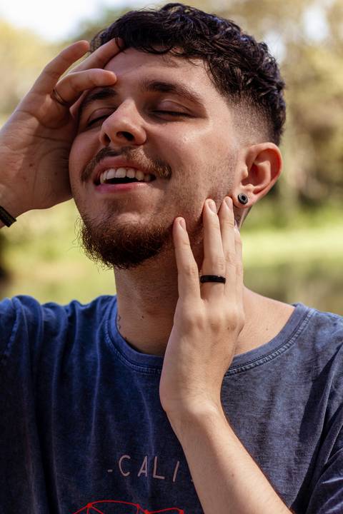 Matheus com árvores ao seu redor no parque do Ibirapuera, na capital de são paulo, em frente ao lago, sorrindo, com as mãos em volta de seu rosto, olhos fechados.'