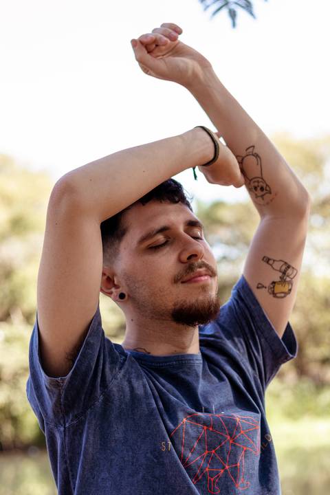 Matheus com árvores ao seu redor no parque do Ibirapuera, na capital de são paulo, em frente ao lago, sorrindo, com as mãos em volta de seu rosto, olhos fechados, sorriso leve.'