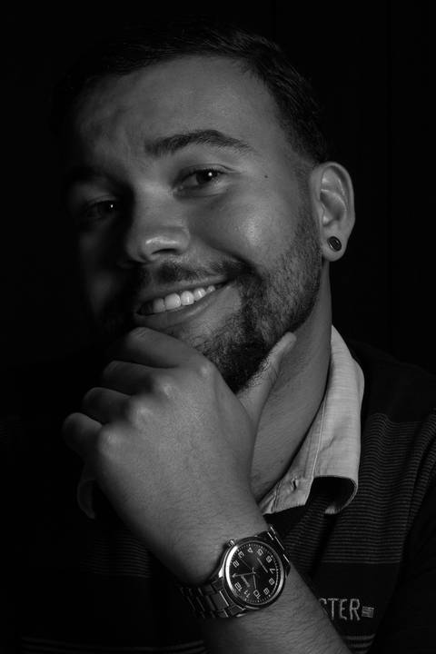 Rafael sorrindo, com mão sobre o queixo, em estúdio fundo preto.'