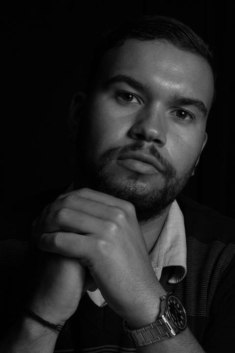 Rafael sério, com as mãos entrelaçadas a frente do queixo, em estúdio com fundo preto.'