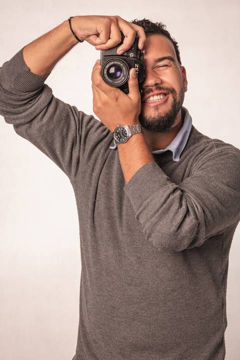 Rafael, em estúdio com fundo branco, segurando uma câmera fotográfica, com o olhar direcionado através do equipamento, como se ele estivesse me fotografando.'