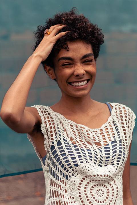 Larissa sorrindo, com a mão em seu cabelo cacheado, fundo de tecido azul marinho.'