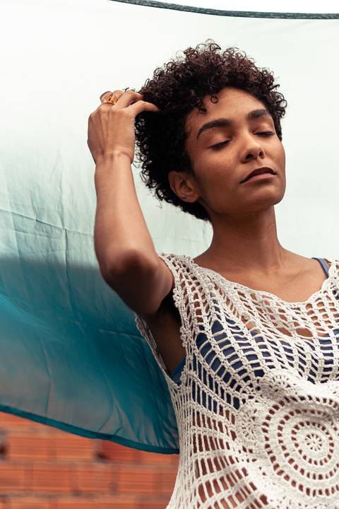 Larissa de olhos fechados, com a mão no cabelo encaracolado, fundo de tecido azul marinho.'