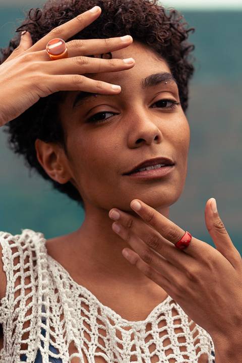 Larissa com olhos atentos a câmera, com as mãos próximas ao rosto, fundo de tecido azul marinho.'