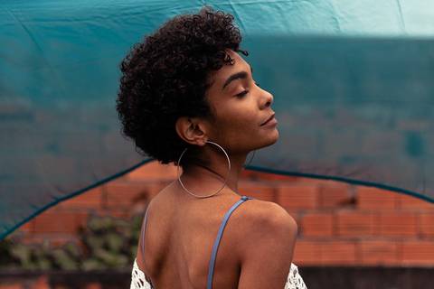 Larissa com os olhos fechados, vento batendo, sorrindo leve, fundo de tecido azul marinho.'