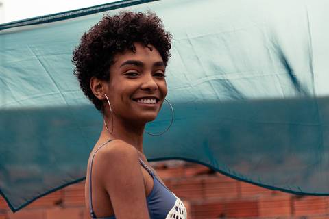 Larissa com o olhar direcionado para a câmera, sorrindo, vento batendo leve, fundo de tecido azul marinho.'
