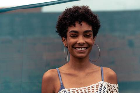 Larissa com o olhar direcionado para a câmera, sorrindo leve, virada para a câmera, fundo de tecido azul marinho.'