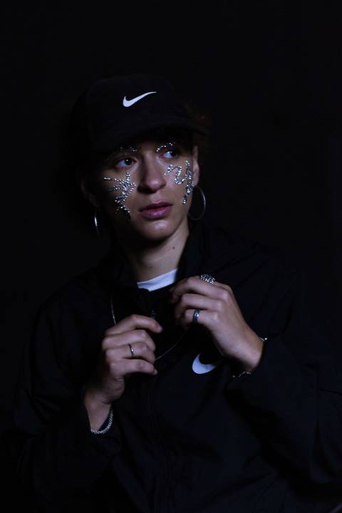 Lariane em cima da laje de uma residência, na zona leste da capital de são paulo. olhando diretamente para mim, repleta de brilhos sob o rosto. rosto bem enquadrado. séria. segurando a blusa da nike.'