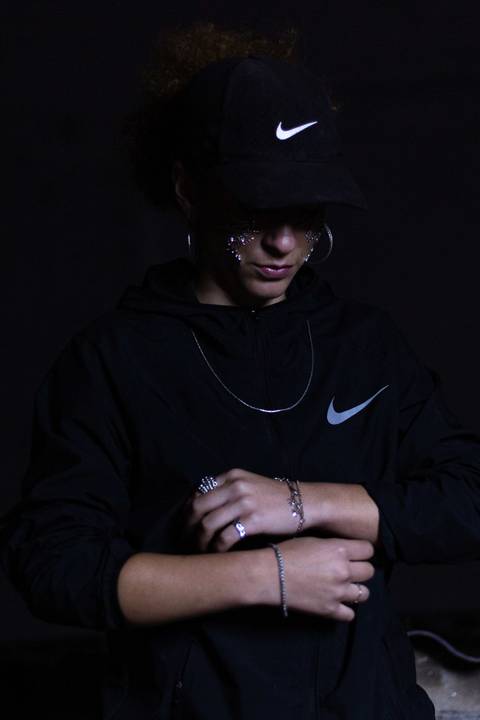 Lariane em cima da laje de uma residência, na zona leste da capital de são paulo. olhando diretamente para mim, repleta de brilhos sob o rosto. rosto bem enquadrado. séria. segurando a blusa da nike. olhando para baixo.'