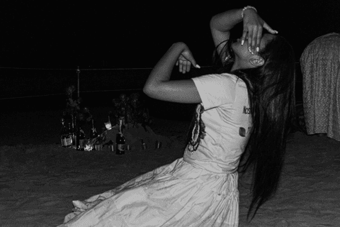 Entidade ajoelhada sob areia com guias alisando seu rosto, cabelos soltos longos. Velas, flores, imagens e bebidas ao fundo.'