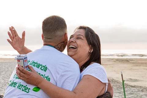 Marinheiros gargalhando em frente ao mar enquanto de abraçam, com latas de cerveja nas mãos.'