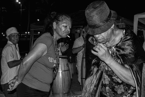Religiosos incorporados conversando em evento religioso umbandista na praia grande, com guias e copo de bebida em mãos. Ao fundo instrumentos de macumba e pessoas tocando.'