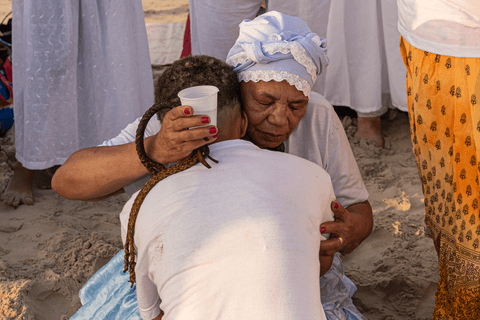 Entidade ajoelhada na areia abraçando outra integrante do terreiro, em momento de passe.'
