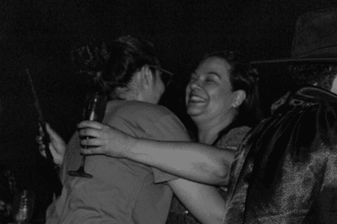 Entidade abraçando fiel e também integrante do terreiro, ambas sorrindo com taça e leque na mão.'