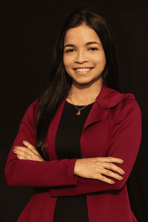 Bruna sorrindo, com seus braços cruzados à frente do corpo, em estúdio com fundo preto.'
