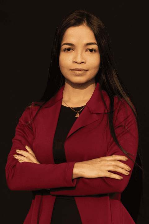 Bruna séria, com seus braços cruzados à frente do corpo, em estúdio com fundo preto.'