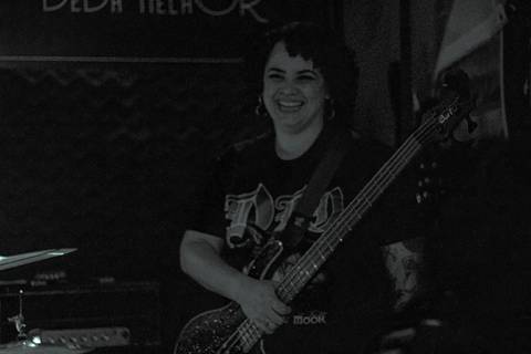 Show da ultra 77 no Armazém 77 aline sorrindo enquanto toca seu baixo, olhando para a câmera'