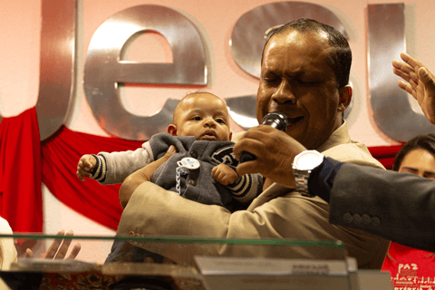 bryan sendo apresentado pelo pastor na frente de toda a igreja, com a palavra jesus escrito ao fundo do palco'