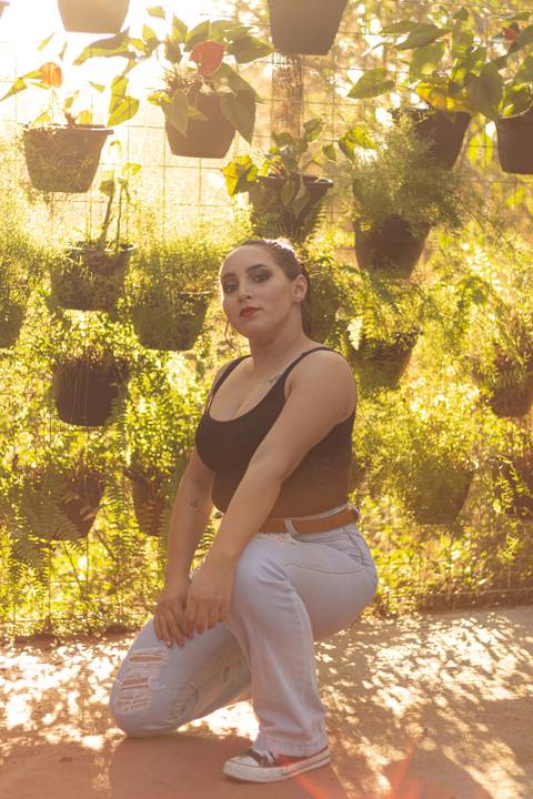 isabel agachada em frente a parede de vasos, dentro da estuda, luz bonita de sol ao fundo'