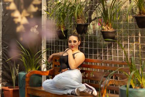 isabel sentada no banco dentro da estufa do parque max feffer corpo inteiro, com flores a frente e vasos no fundo'