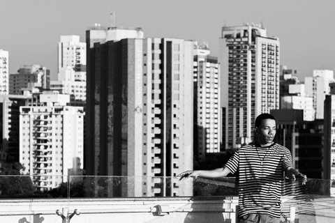 jerf no terraço do mac em pé segurando o corrimão, prédios da cidade ao fundo, olhando para a lateral, foto em preto e branco'