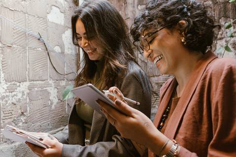 le e ni em pé, ambas sorrindo enquanto analisam seus arquivos e documentos de trabalho'