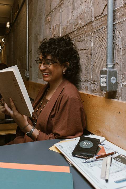 leticia sentada a mesa da cafeteria lendo um livro de arquitetura enquanto sorri'