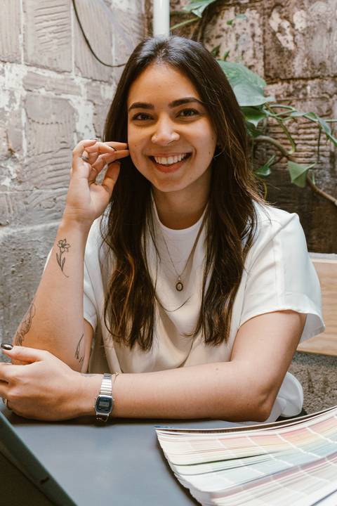 nicole com o braço apoiado na mesa, olhando para mim, sorrindo com dentes a mostra, mão ao lado do rosto'