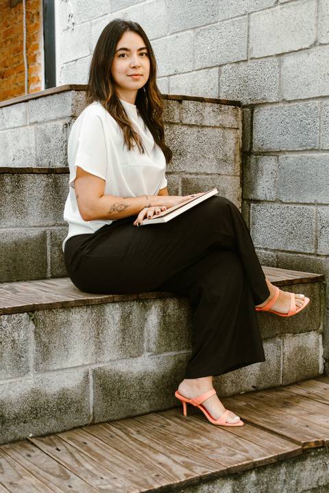 nicole na parte de fora da cafeteria, próximo a escadaria, sentada com as pernas cruzadas e livro de arquitetura sobre o colo'