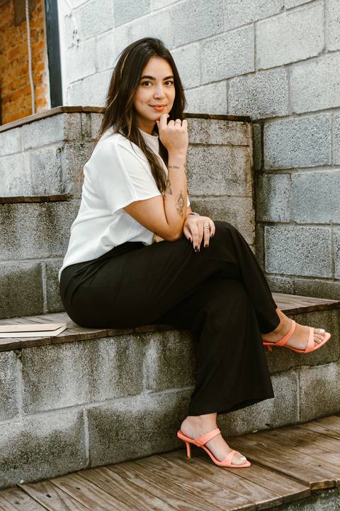 nicole na parte de fora da cafeteria, próximo a escadaria, sentada, com o braço apoiado em livro em seu colo, sorrindo'