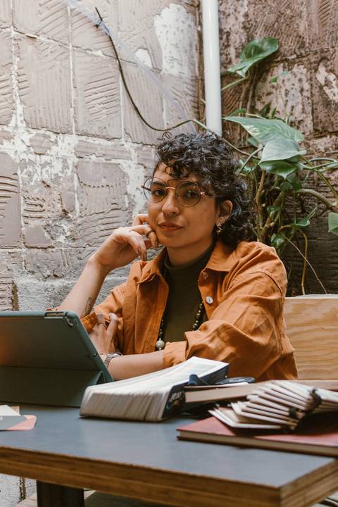 leticia sentada dentro da cafeteria, com o tablet, braço apoiado na mesa enquanto mão toca seu rosto'