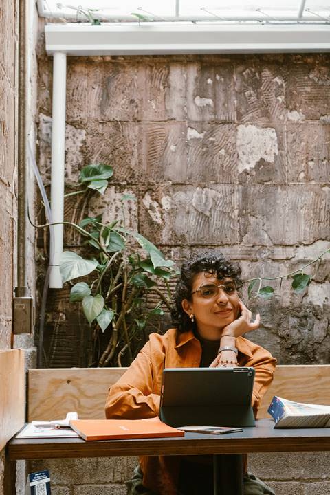 leticia sentada dentro da cafeteria, com o tablet, com o rosto apoiado em sua mão, plantas ao fundo'