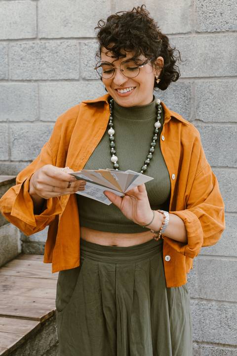 leticia na parte de fora da cafeteria em frente a escadaria, segurando paleta de cores nas mãos enquanto sorri'