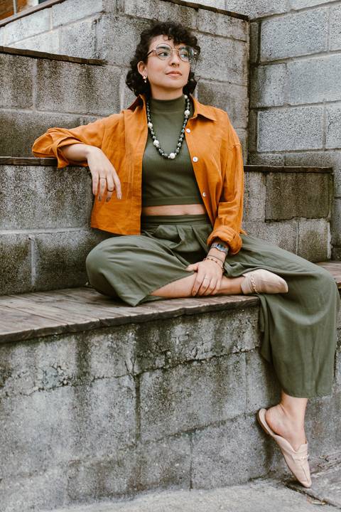 leticia sentada na escadaria em frente a cafeteria, com uma das pernas cruzadas, enquanto um dos braços apoia na escadaria também'