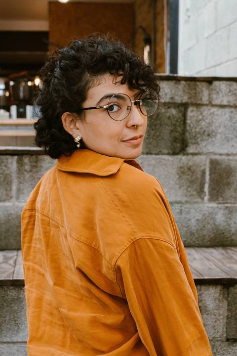 leticia em pé na escadaria em frente a cafeteria,  olhando por cima dos ombros, de costas'