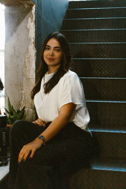 nicole sentada na escadaria na parte de dentro da cafeteria, olhando para a frente, foto em colorido'