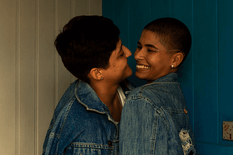 mika e le em enquadramento mais fechado, na parte interna da cafeteria, quase se beijando, sorrindo'