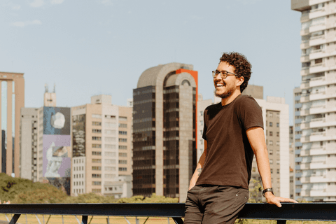 Leo no centro cultural são paulo, em pé escontado no corrimão, enquanto apoia suas mãos nele, sorrindo, com os prédios de fundo no cenário'