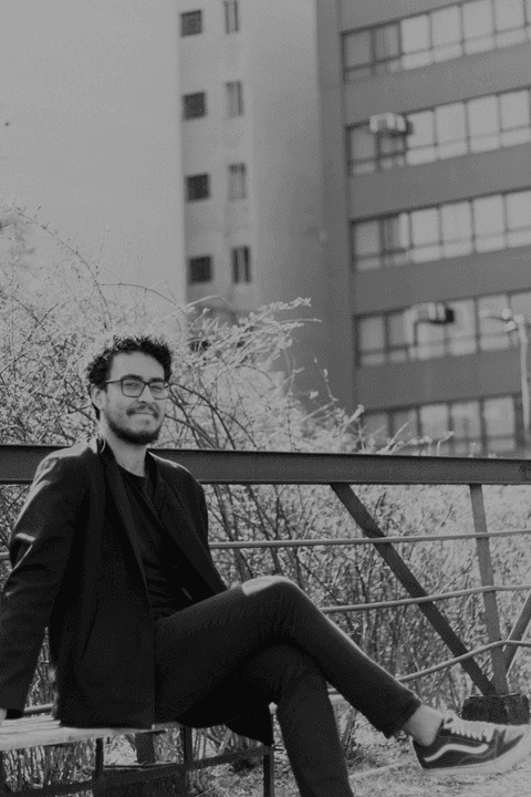 Leo no centro cultural são paulo, sentado no banco, sorrindo, com a arquitetura dos prédios do centro ao fundo, foto em preto e branco'
