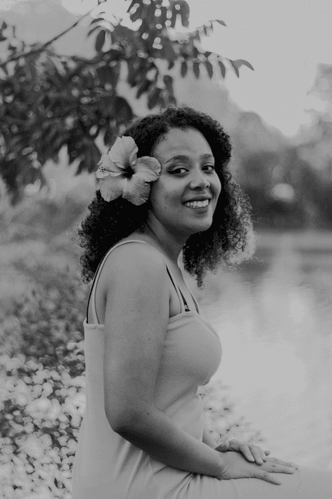 leticia meio corpo com a flor rosa atrás da orelha, sentada em frente ao lago, sorrindo, foto em preto e branco'