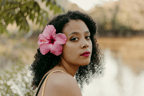 leticia meio corpo com a flor rosa atrás da orelha, sentada em frente ao lago, mais séria, olhando em direção a câmera'