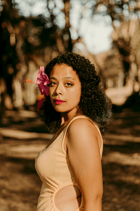 lety olhando para mim, com seu cabelo solto, flor rosa atrás da orelha, corpo de lateral'